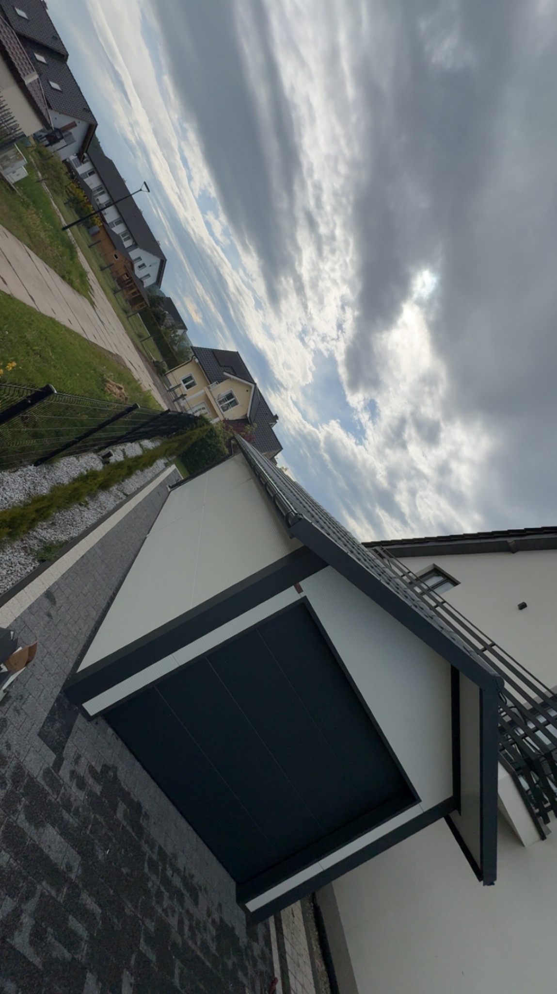 Nowoczesny garaż blaszany z bramą segmentową w kolorze antracytowym, na tle domu jednorodzinnego i pochmurnego nieba. Widok z dołu, podkreślający solidną konstrukcję.