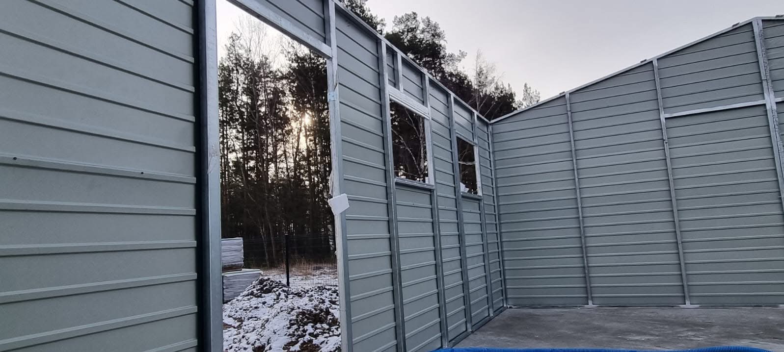 Montaż szarego garażu blaszanego z widocznymi oknami i ramą drzwiową. W tle drzewa i resztki śniegu. Widoczny etap budowy.