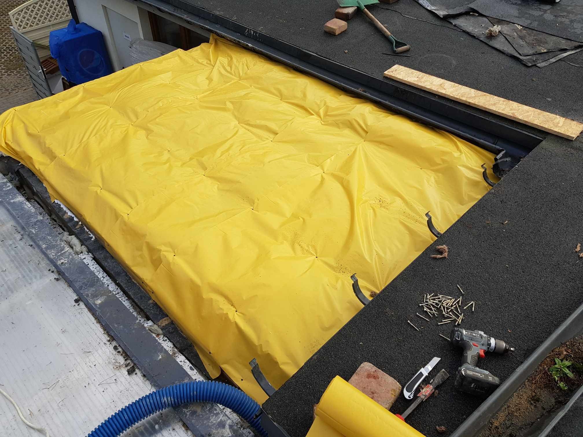 Prace na dachu: żółta folia ochronna rozłożona na fragmencie dachu, obok wkrętarka, wkręty, nóż i fragment rynny z ziemią i roślinnością.