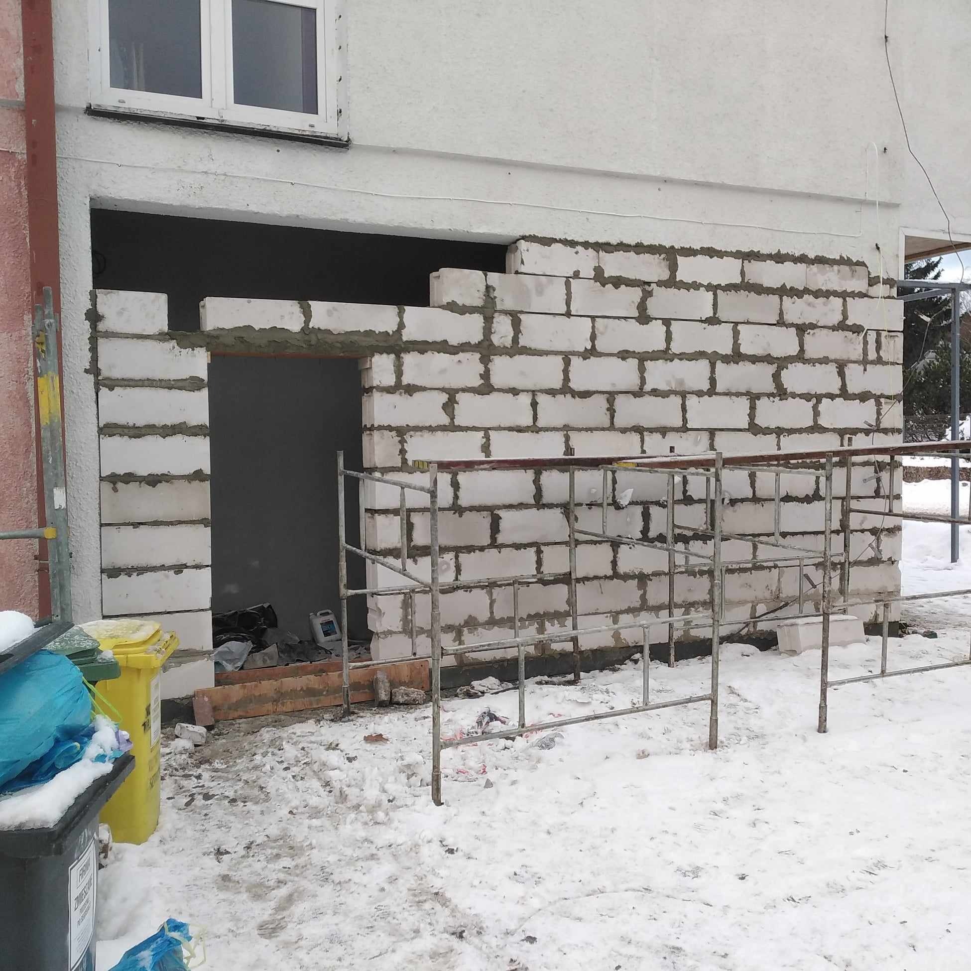 Wznoszenie ściany z białych bloczków betonowych z pozostawionym otworem na drzwi w Koszalinie, widoczne rusztowanie, ślady zaprawy i śnieg na ziemi.