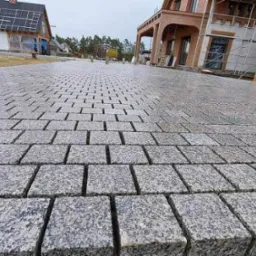 Niedokończona nawierzchnia z kostki granitowej w odcieniach szarości, ułożona w równych rzędach, z widocznym podsypem piaskowym i fragmentami budynków w tle.