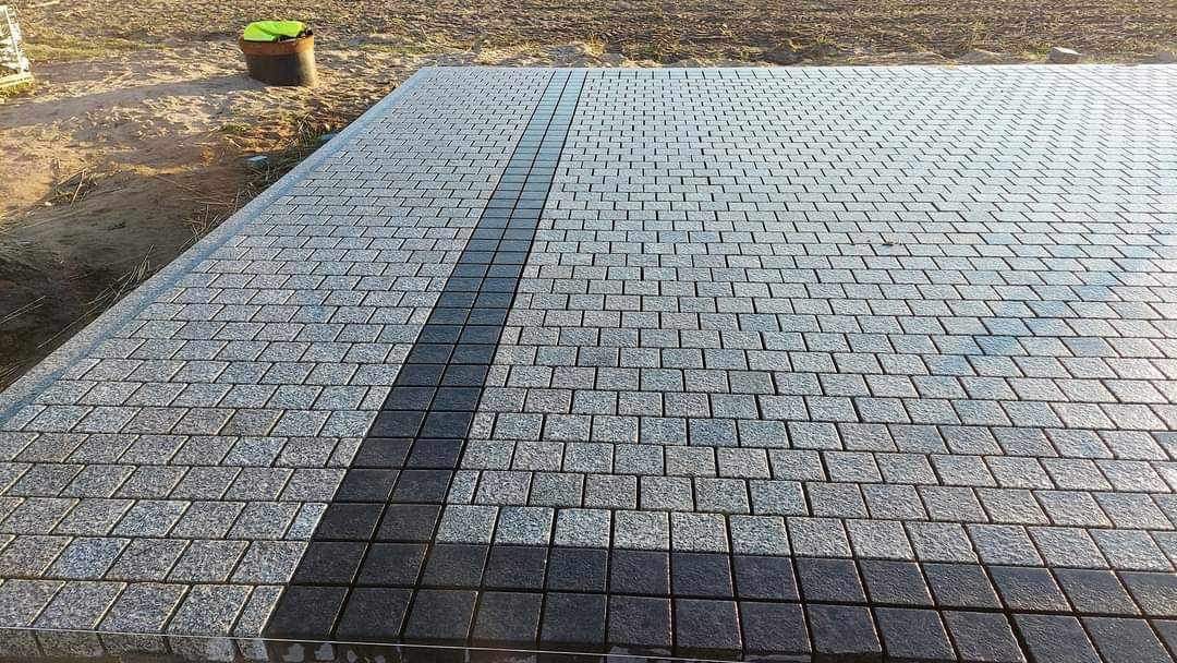 Świeżo ułożona nawierzchnia z kostki granitowej w odcieniach szarości, z ciemniejszym pasem biegnącym przez środek, na tle ziemi i wiadra.