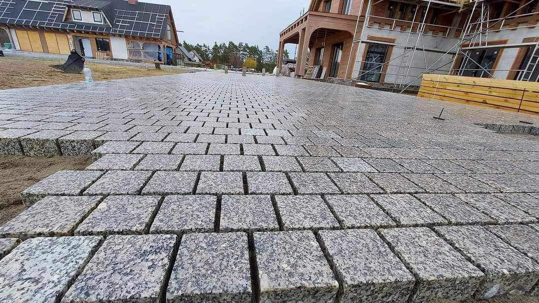 Niedokończona nawierzchnia z kostki granitowej w odcieniach szarości, ułożona w równych rzędach, z widocznym podsypem piaskowym i fragmentami budynków w tle.