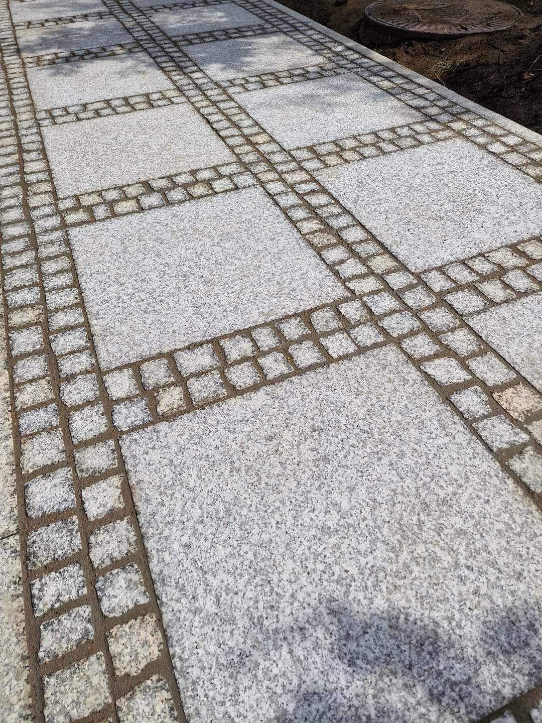 Ułożona nawierzchnia z jasnoszarej kostki granitowej, z kwadratowymi panelami obramowanymi mniejszą kostką, widok ukośny.