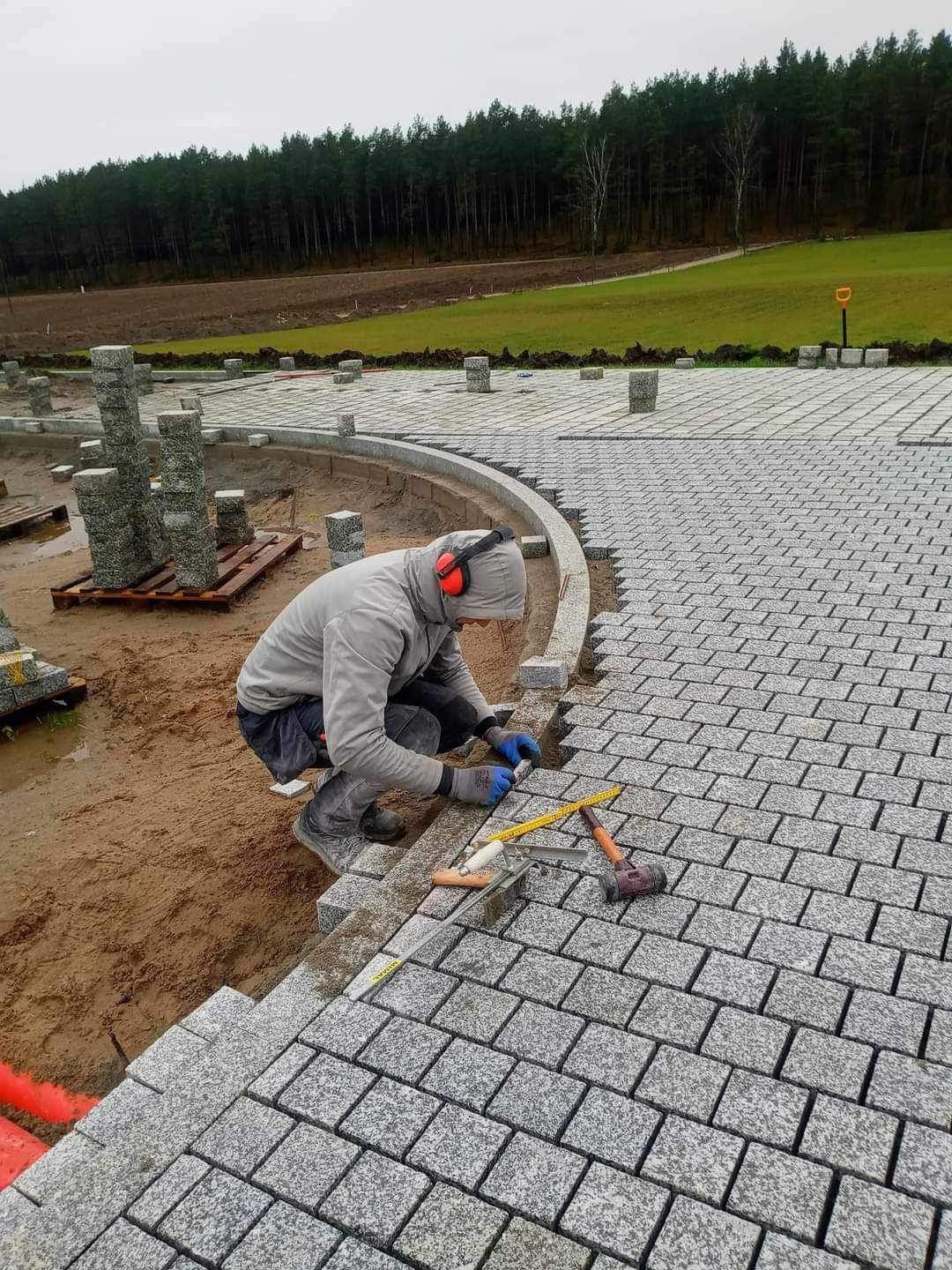 Pracownik w szarej bluzie i słuchawkach ochronnych układa kostkę granitową na podsypce, widoczne narzędzia: młotek, poziomica, łata, w tle teren zielony z drzewami i ułożone słupki z kostki.