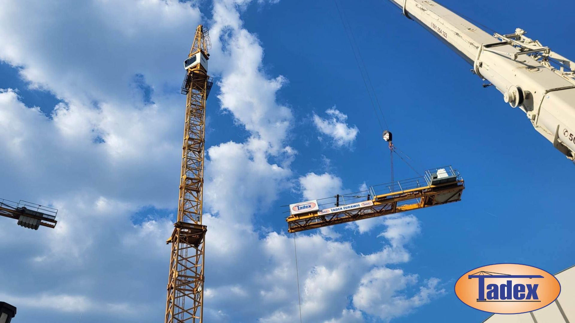 Żuraw wieżowy w trakcie montażu platformy roboczej, widoczne logo Tadex na platformie, błękitne niebo z chmurami w tle.