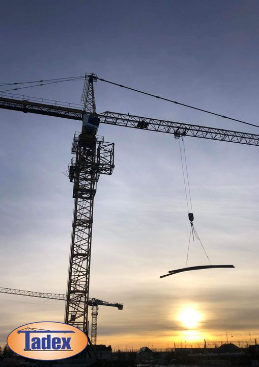 Żuraw wieżowy w trakcie podnoszenia elementu konstrukcyjnego na tle zachodzącego słońca, widoczny budynek w tle i logo firmy Tadex.
