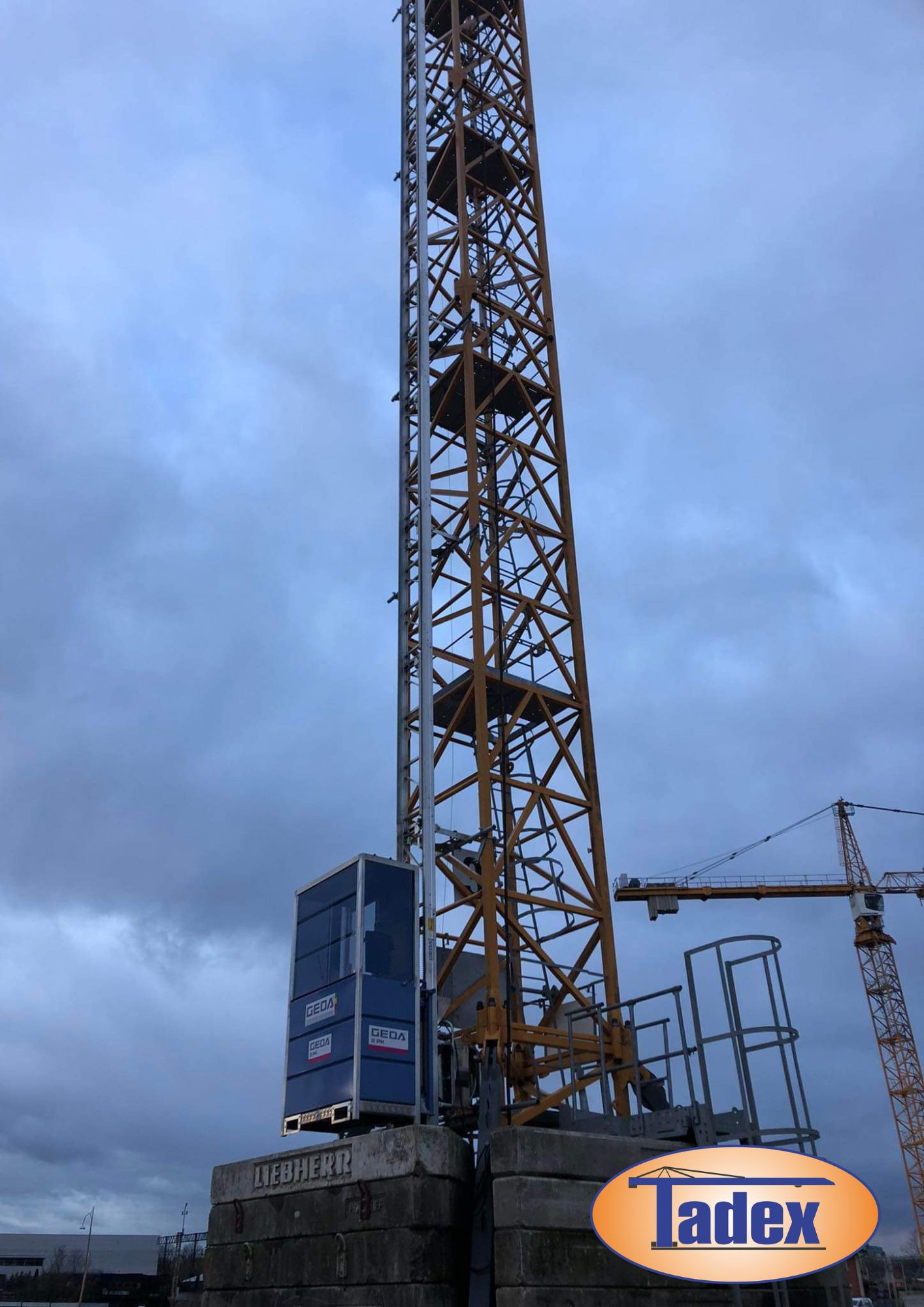 Wysoki żuraw wieżowy z windą osobową GEDA, widoczny na tle pochmurnego nieba, z drugim żurawiem w tle po prawej stronie.