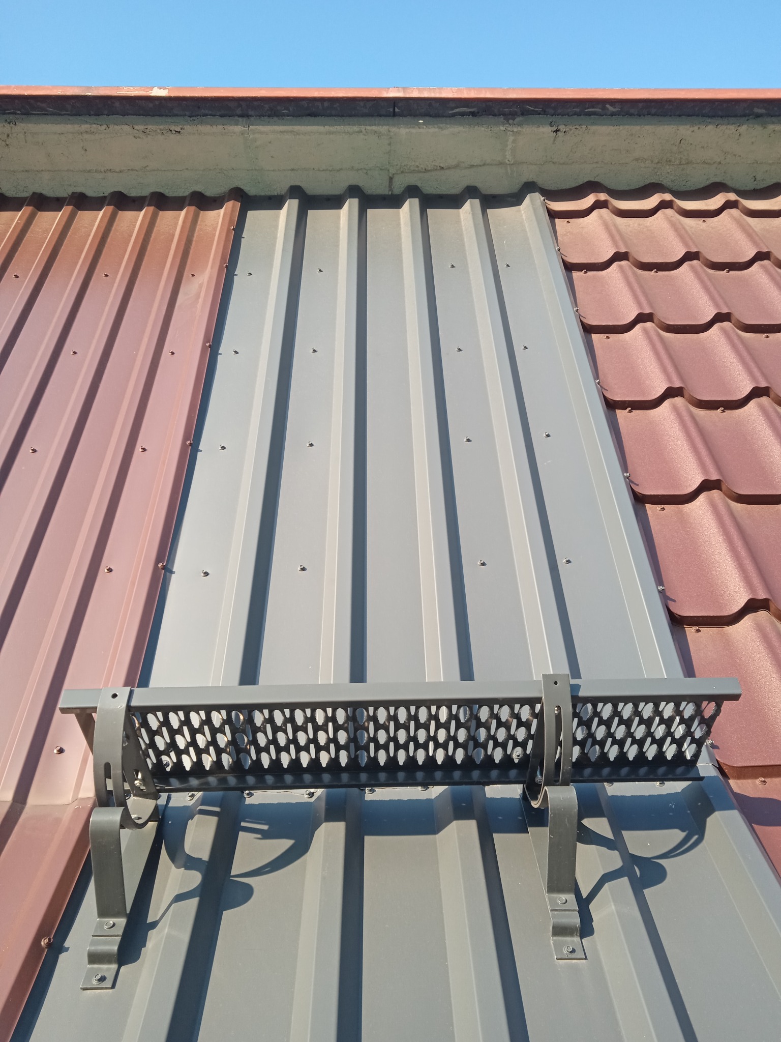 Fragment dachu z blachy trapezowej w kolorze szarym, brązowym i dachówką ceramiczną w kolorze czerwonym, z zamontowanym zabezpieczeniem przeciwśniegowym.