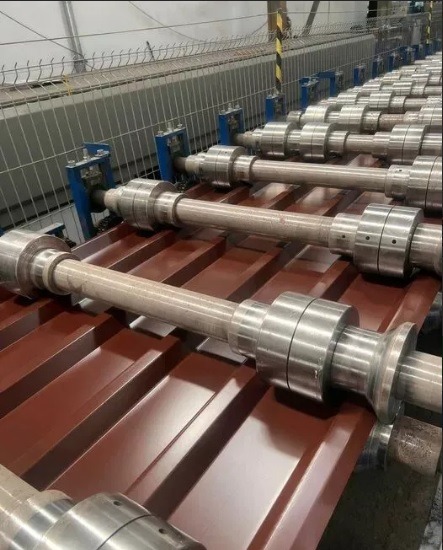 Proces formowania blachy trapezowej na linii produkcyjnej z użyciem walców profilujących. Widoczne metalowe rolki i uformowany, brązowy arkusz blachy.