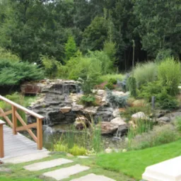 Ogród z drewnianym mostkiem nad stawem i kaskadą wodną, otoczony różnorodną roślinnością, w tle gęsty las.