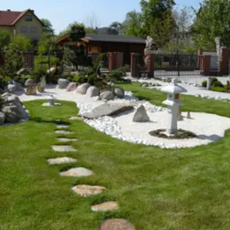 Ogród w stylu japońskim z kamienną ścieżką, białymi kamykami, ozdobną latarnią i zadbaną zielenią na tle drewnianego domku i murowanego ogrodzenia.