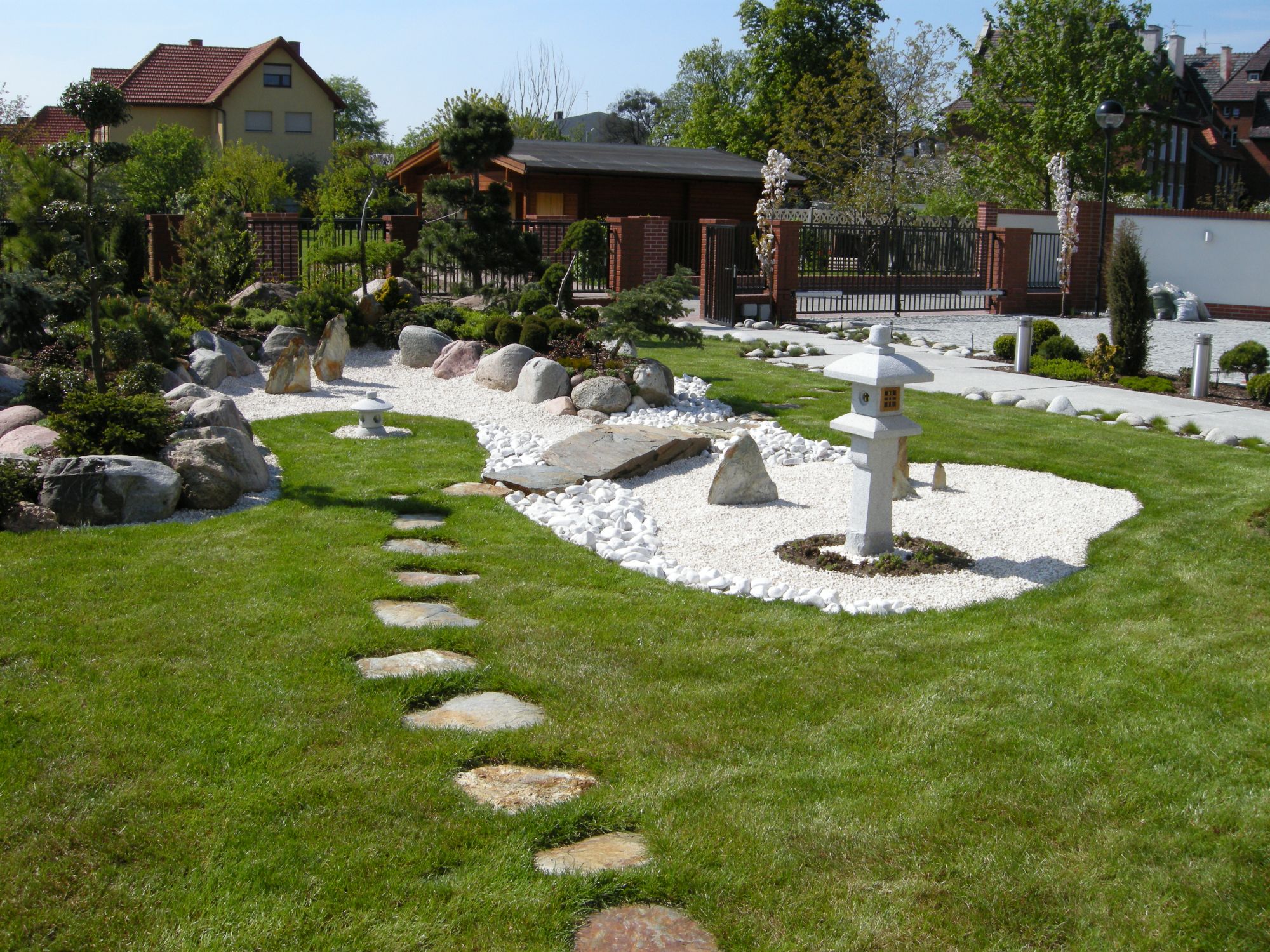 Ogród w stylu japońskim z kamienną ścieżką, białymi kamykami, ozdobną latarnią i zadbaną zielenią na tle drewnianego domku i murowanego ogrodzenia.