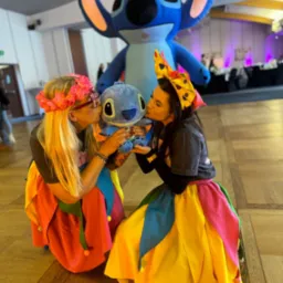 Dwie kobiety w kolorowych strojach klęczą i całują pluszową zabawkę Stitch, w tle dmuchana postać Stitch na sali eventowej z dekoracjami.
