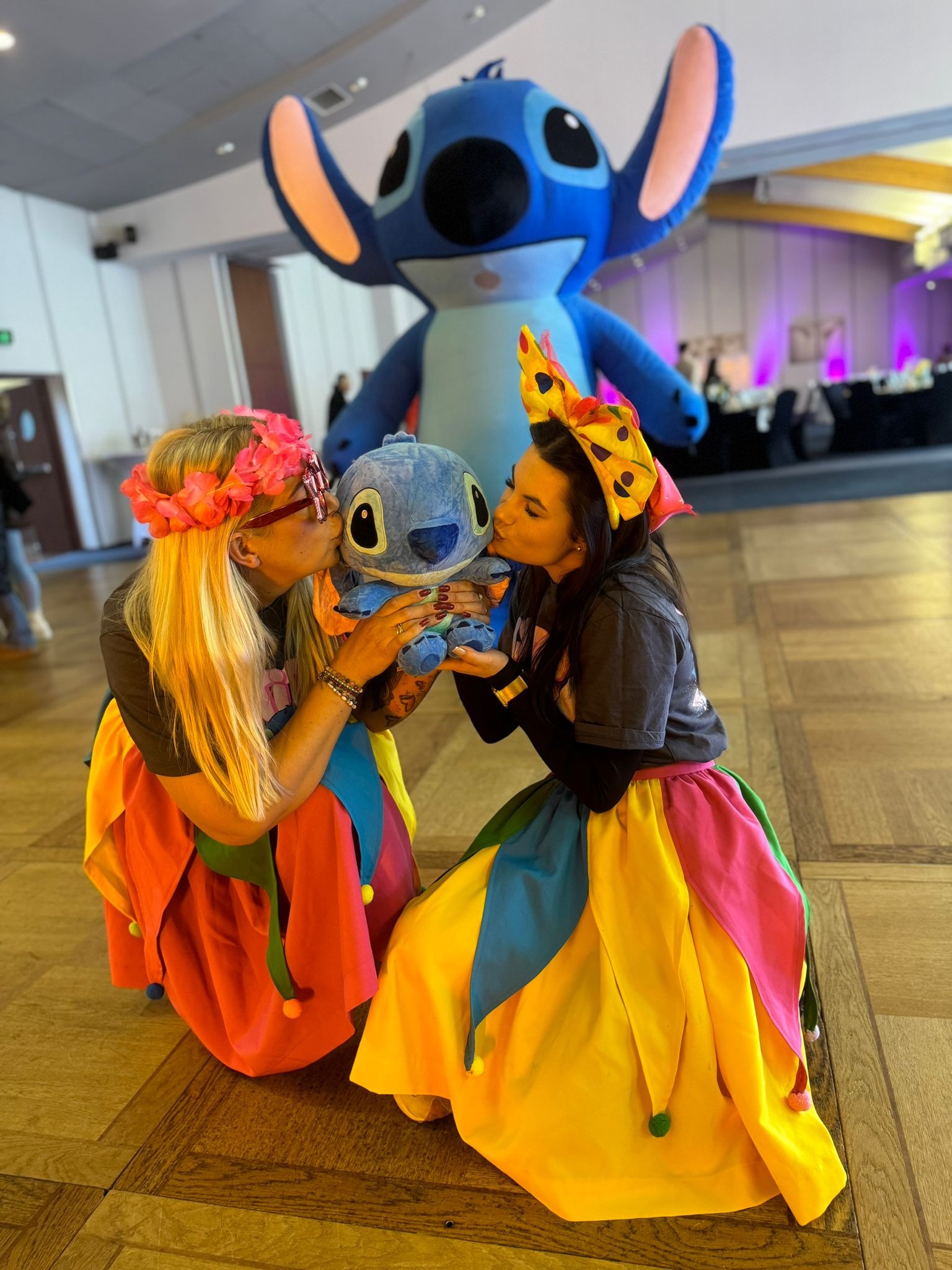 Dwie kobiety w kolorowych strojach klęczą i całują pluszową zabawkę Stitch, w tle dmuchana postać Stitch na sali eventowej z dekoracjami.