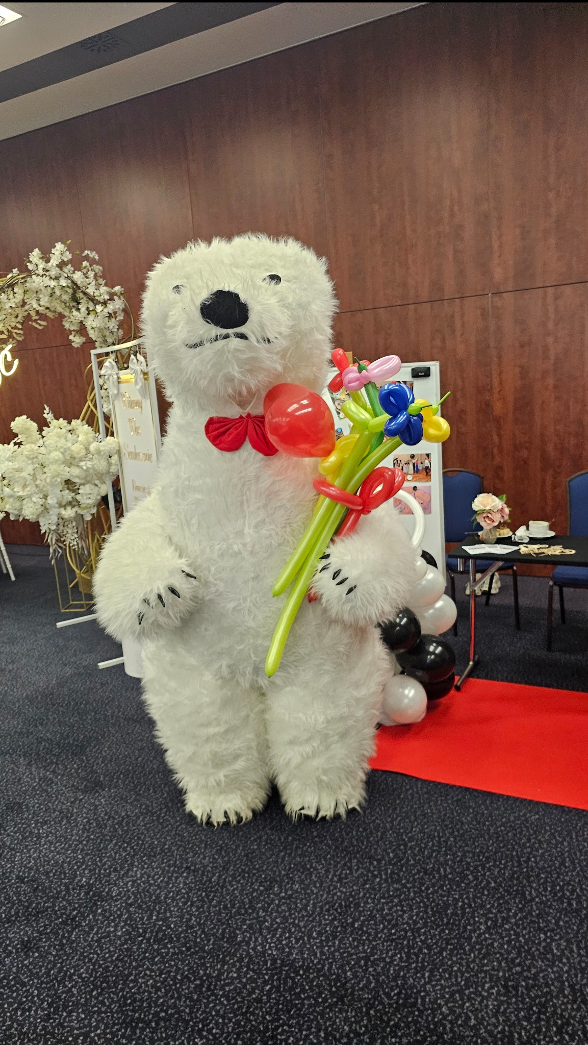 Pluszowy biały miś w czerwonej muszce trzyma bukiet balonów na tle dekoracji eventowej.