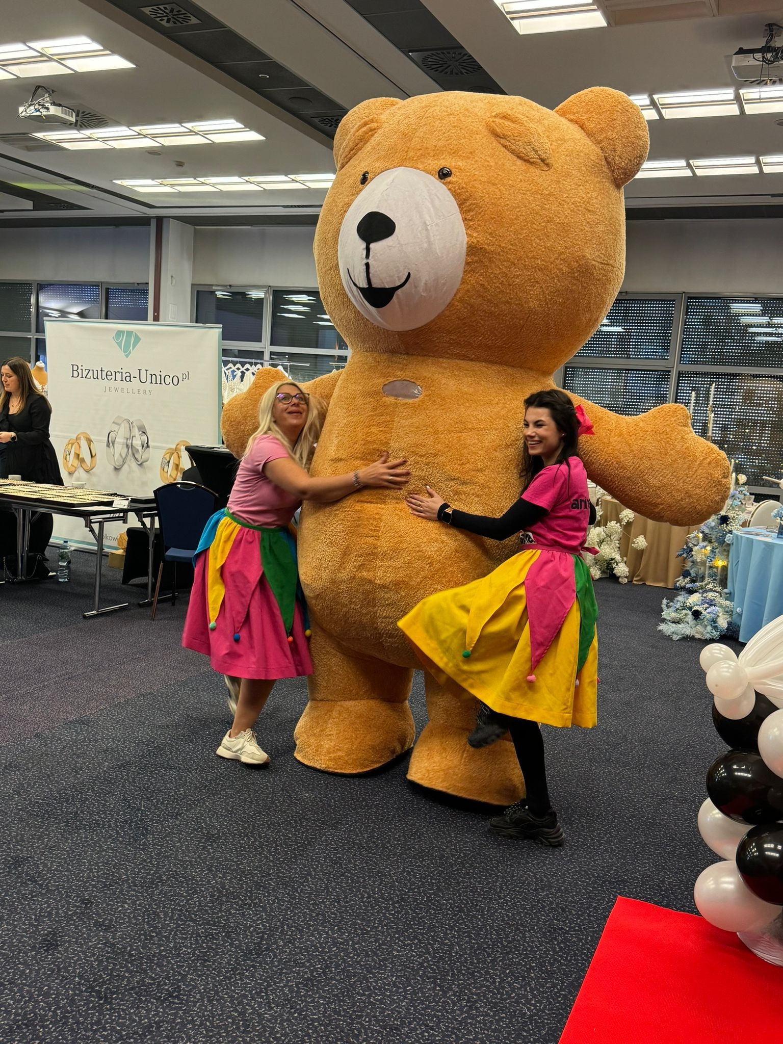 Dwie uśmiechnięte kobiety w kolorowych strojach obejmują dużą maskotkę pluszowego misia na tle sali eventowej z dekoracjami.