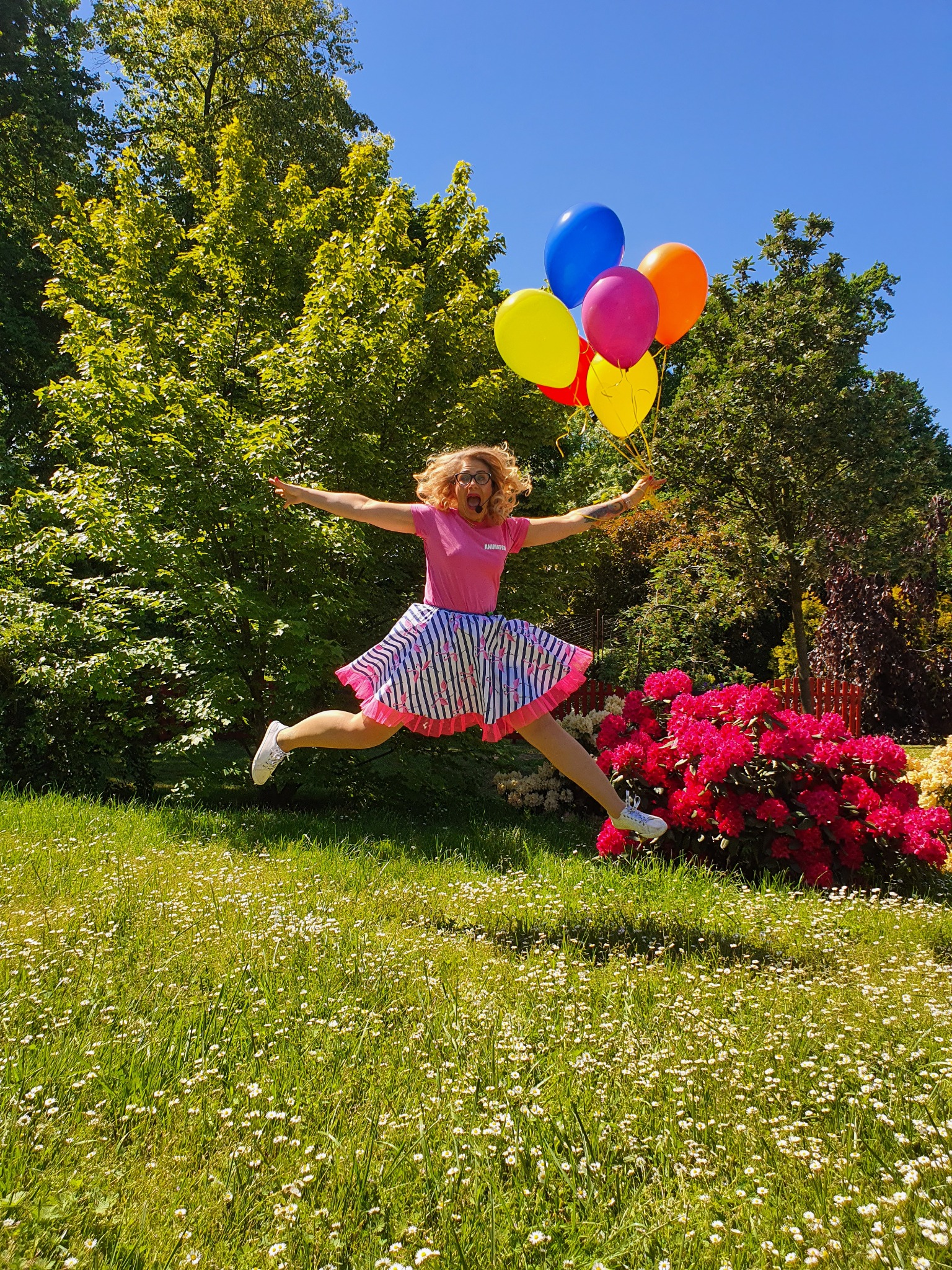 Kobieta w różowej koszulce i spódnicy w paski skacze na trawie, trzymając w ręku kolorowe balony; w tle drzewa i krzew rododendronu.