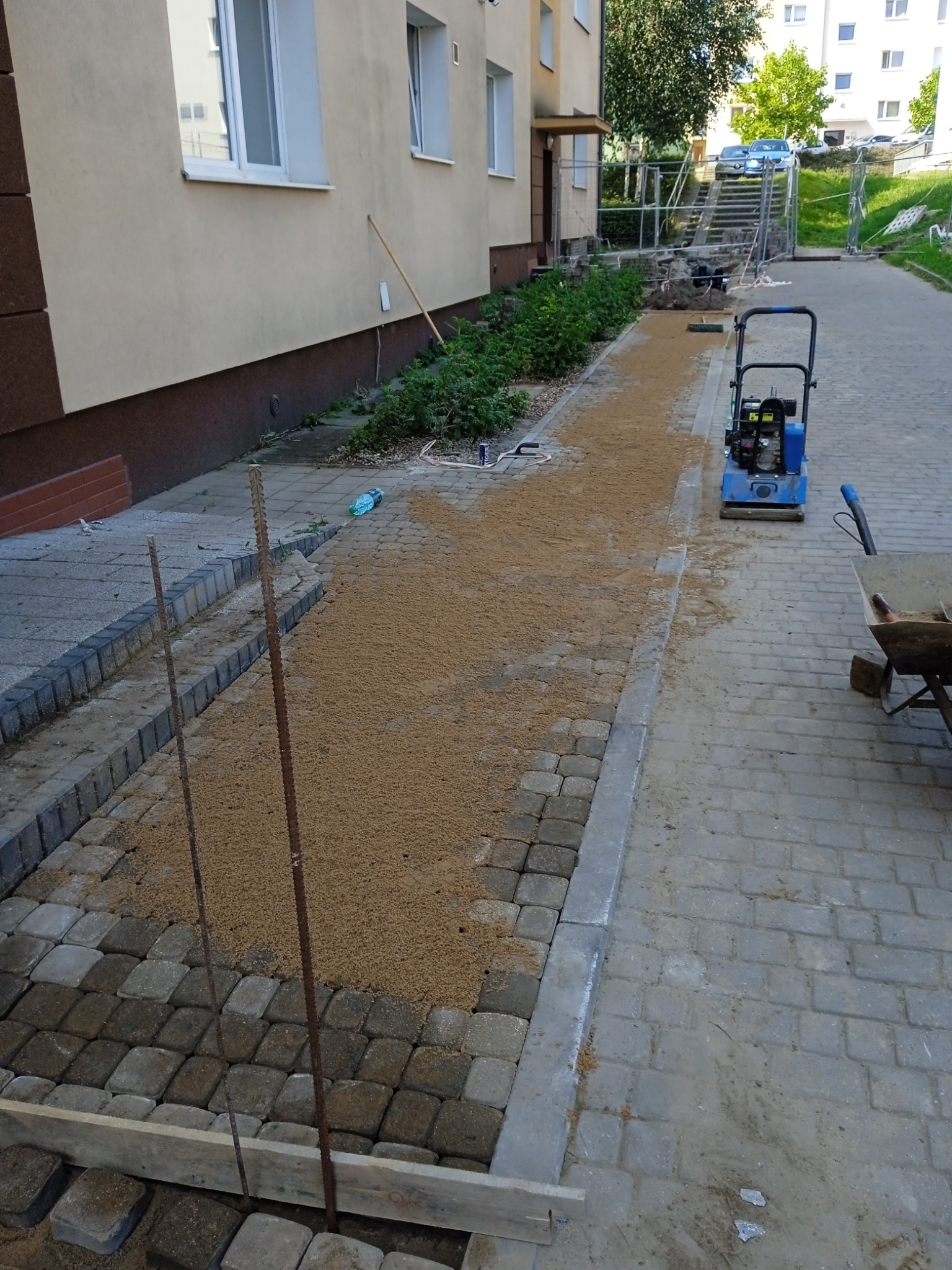 Układanie kostki brukowej na ścieżce wzdłuż budynku, widoczna ubijarka i piasek podsypkowy.