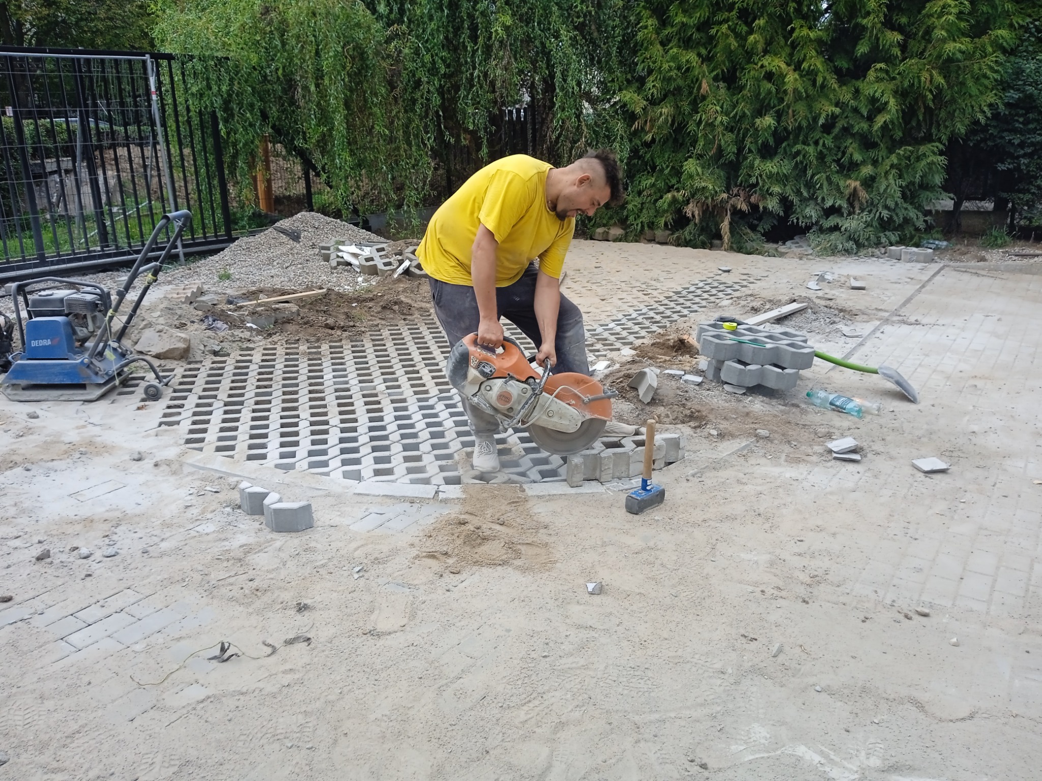 Pracownik w żółtej koszulce tnie betonową kostkę brukową za pomocą piły spalinowej Stihl na piaszczystym podłożu, obok leżą fragmenty kostki, gumowy młotek i maszyna do ubijania gruntu.