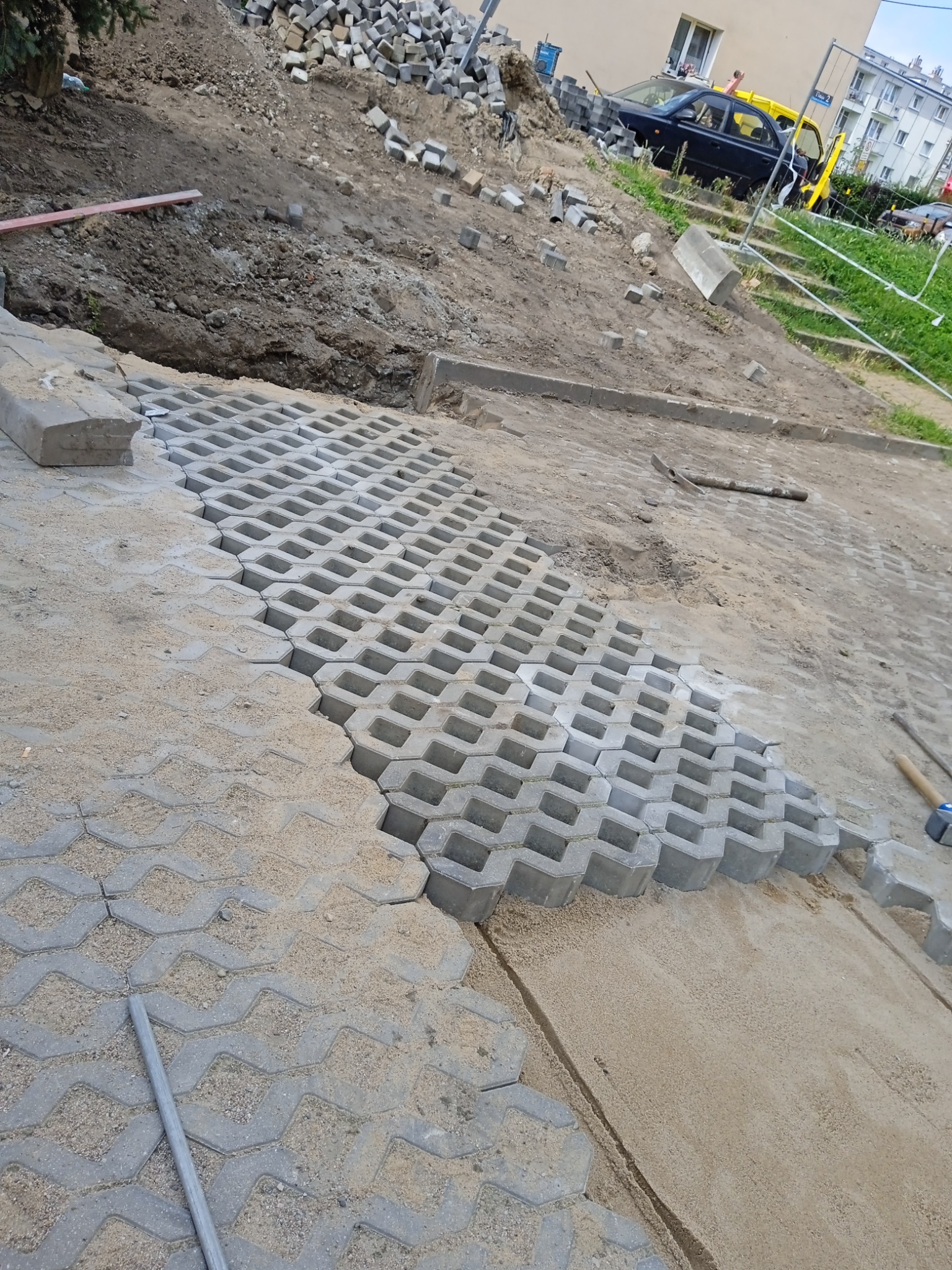 Układanie ażurowych płyt betonowych na podsypce piaskowej, widoczne narzędzia brukarskie i hałda kostki brukowej w tle.