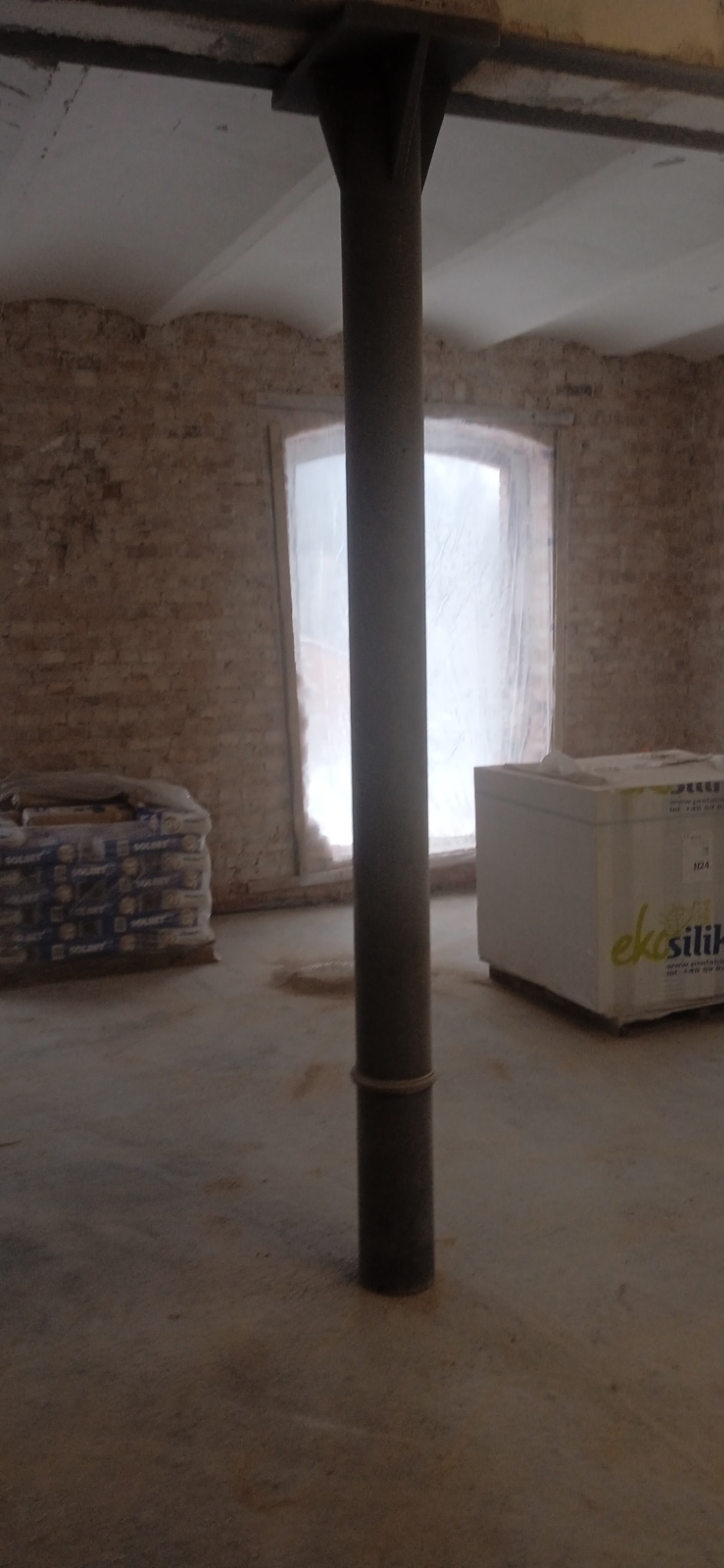 Wnętrze w trakcie remontu w Starogardzie Gdańskim: widoczna ceglana ściana, kolumna nośna, okno zabezpieczone folią, worki z zaprawą i kartony z materiałami budowlanymi.