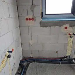 Surowy stan instalacji w budynku z widocznymi przewodami elektrycznymi, rurami wodnymi i kanalizacyjnymi, zabezpieczonymi pianką montażową, oraz puszkami elektrycznymi w ścianie z betonu...