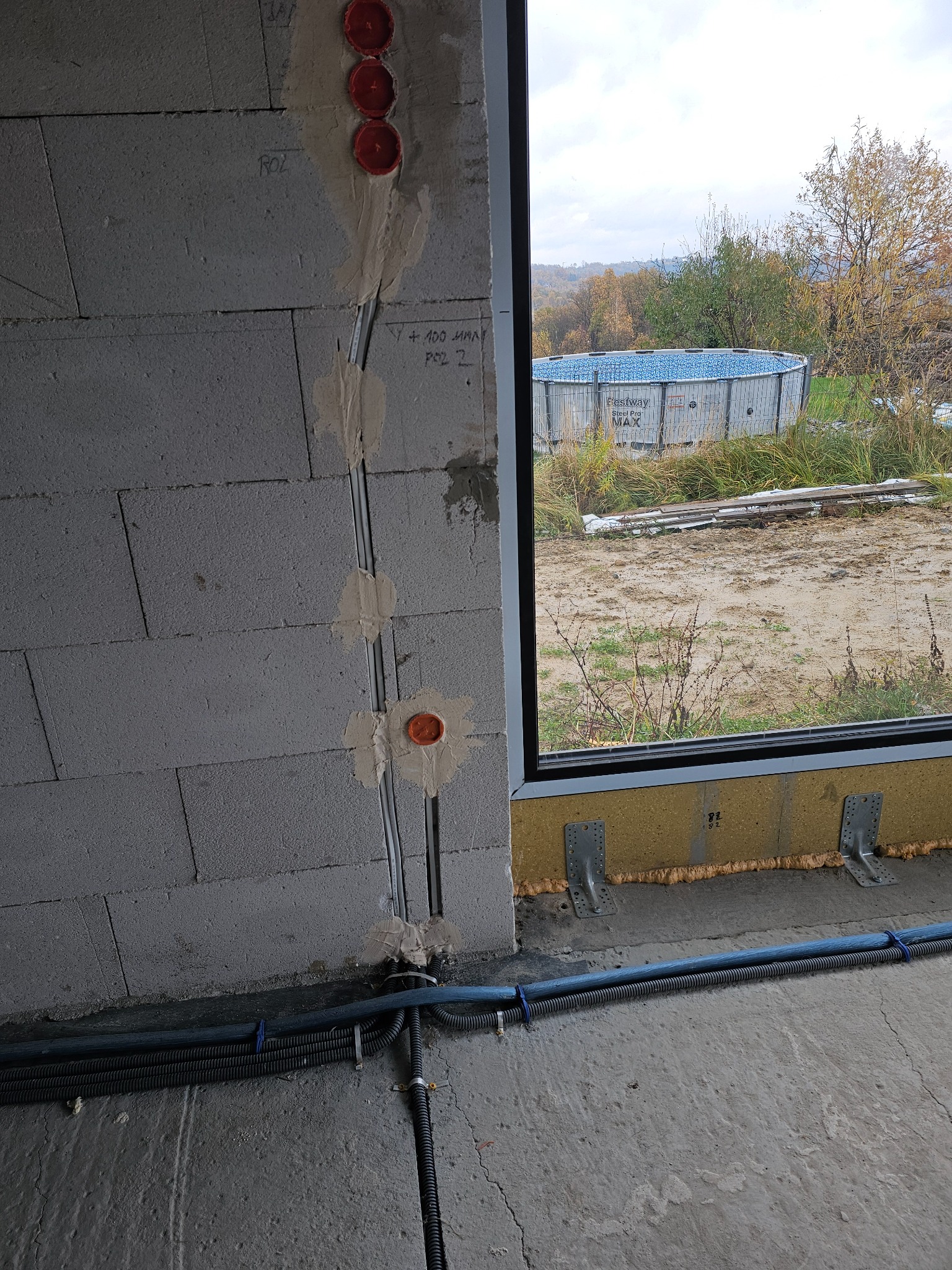 Surowy mur z betonu komórkowego z pionowo ułożonymi peszlami elektrycznymi, widoczne puszki instalacyjne i przewody, w tle przez okno widok na ogród i basen Bestway Steel Pro MAX.