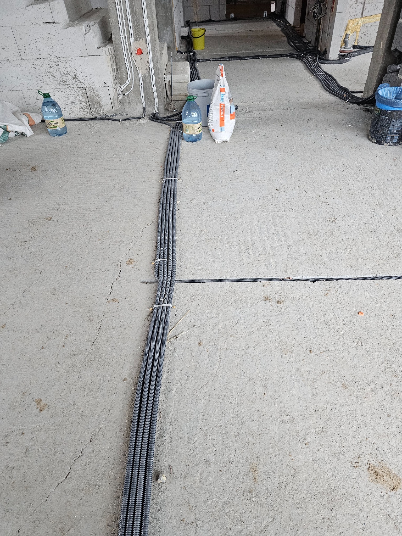 Kable elektryczne w peszlach ułożone na surowym betonie w trakcie montażu, z widocznymi puszkami instalacyjnymi w ścianie i workiem zaprawy murarskiej w tle.