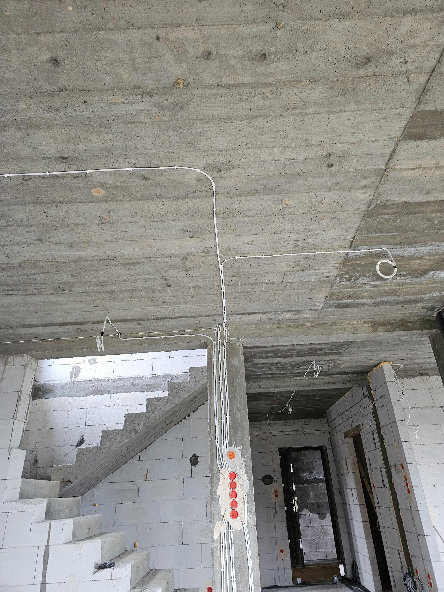 Surowy betonowy strop z poprowadzonymi białymi przewodami elektrycznymi, widoczne puszki instalacyjne, ściany z białych bloczków, w tle schody i otwór drzwiowy.