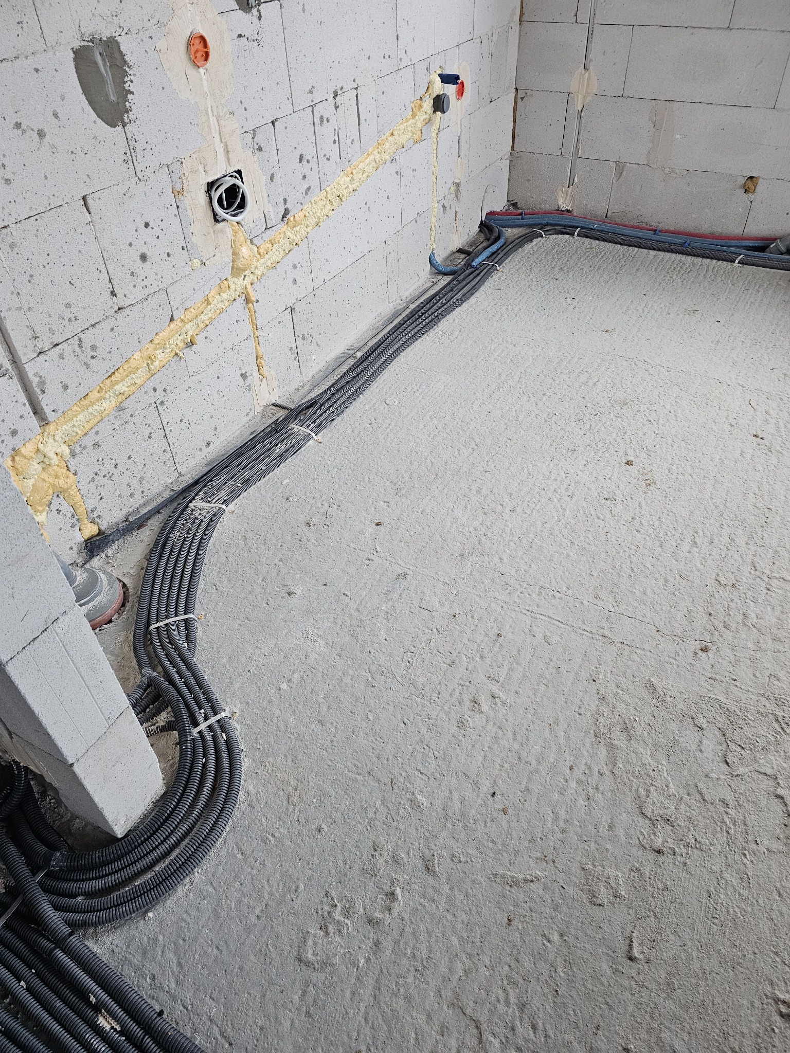 Niedokończona instalacja elektryczna w surowym wnętrzu: wiązki czarnych kabli w peszlach ułożone na betonowej posadzce, prowadzone wzdłuż ściany z betonu komórkowego, zabezpieczone pianką montażową...