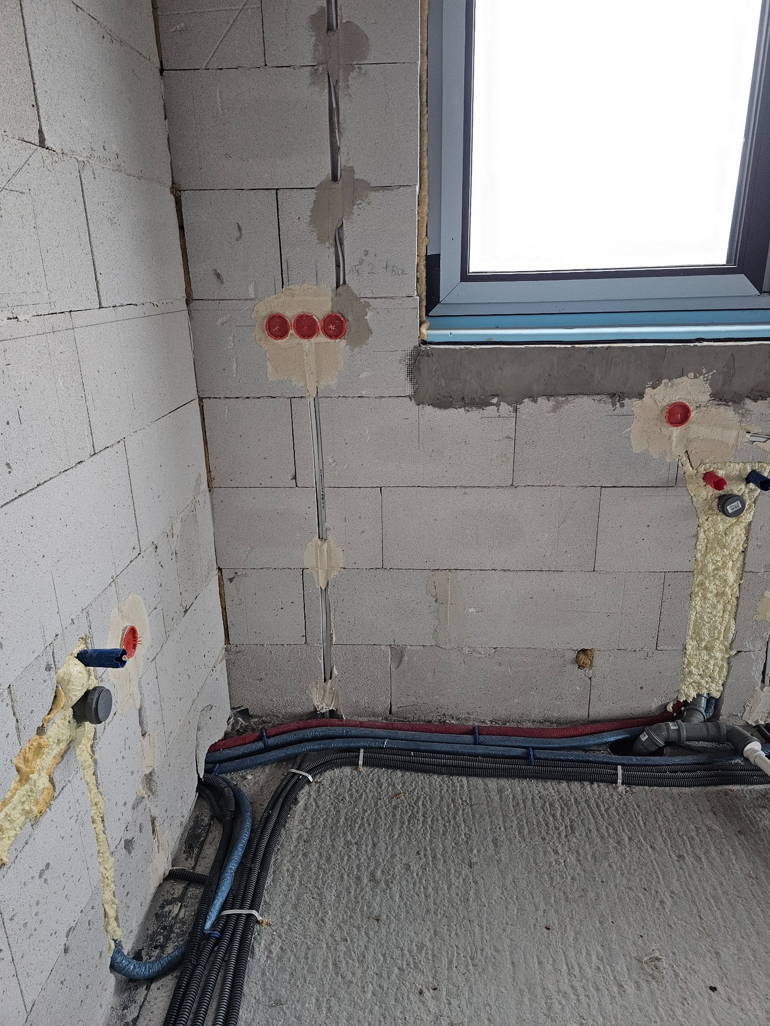 Surowy stan instalacji w budynku z widocznymi przewodami elektrycznymi, rurami wodnymi i kanalizacyjnymi, zabezpieczonymi pianką montażową, oraz puszkami elektrycznymi w ścianie z betonu...