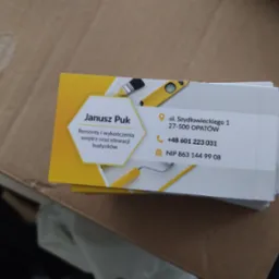 Stos wizytówek firmy remontowo-wykończeniowej Janusz Puk, z adresem i numerem telefonu, leżący na tekturowym tle.