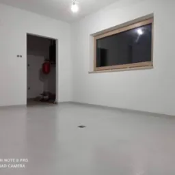 Wnętrze świeżo wykończonego pokoju z gładkimi, białymi ścianami, drewnianym oknem i szarą wylewką podłogową. Widoczny grzejnik w pomieszczeniu obok przez otwarte drzwi.