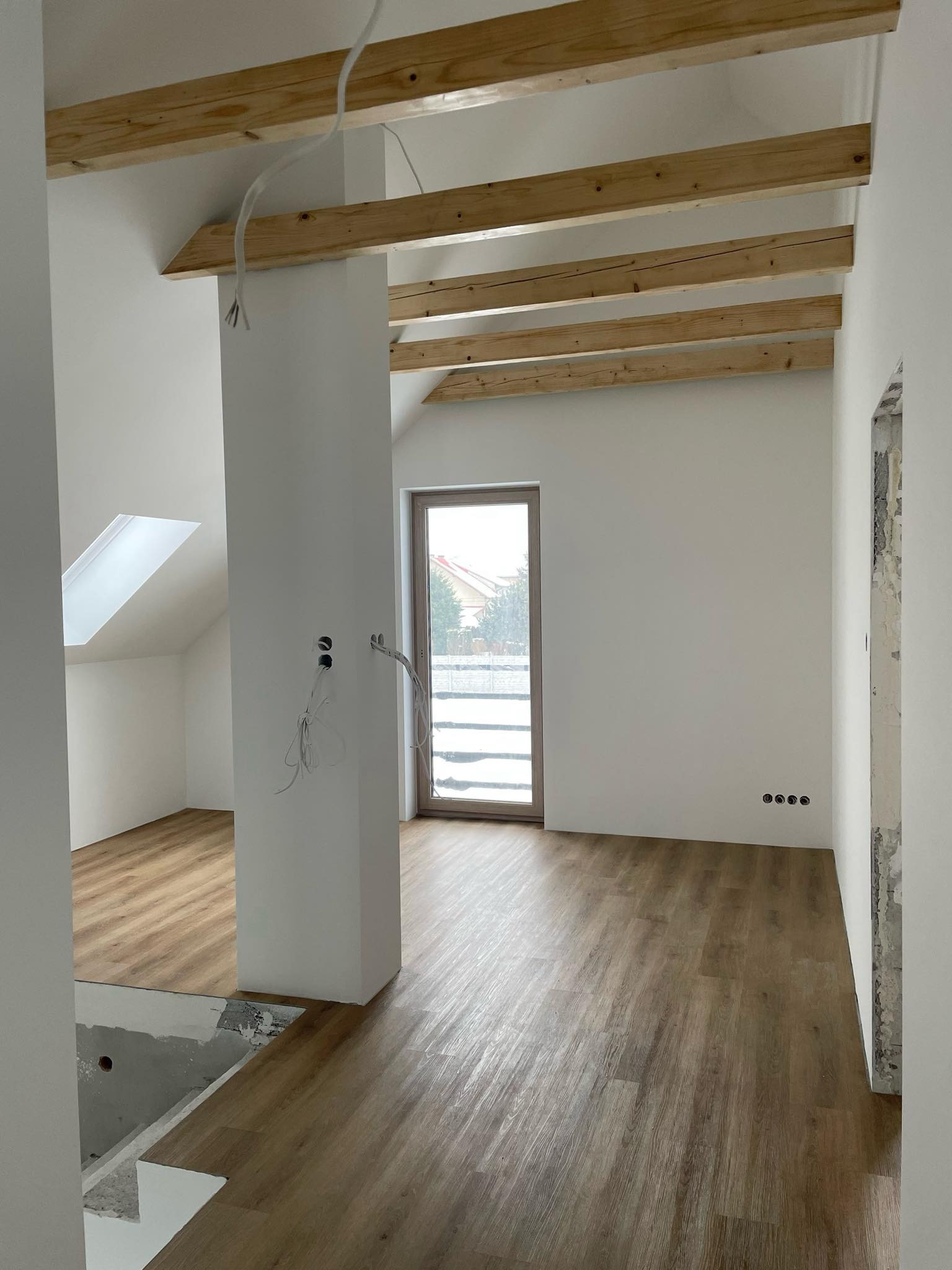 Wnętrze w trakcie remontu z drewnianą podłogą, odsłoniętymi belkami stropowymi i instalacją elektryczną, widoczne gniazdka elektryczne i okno dachowe.