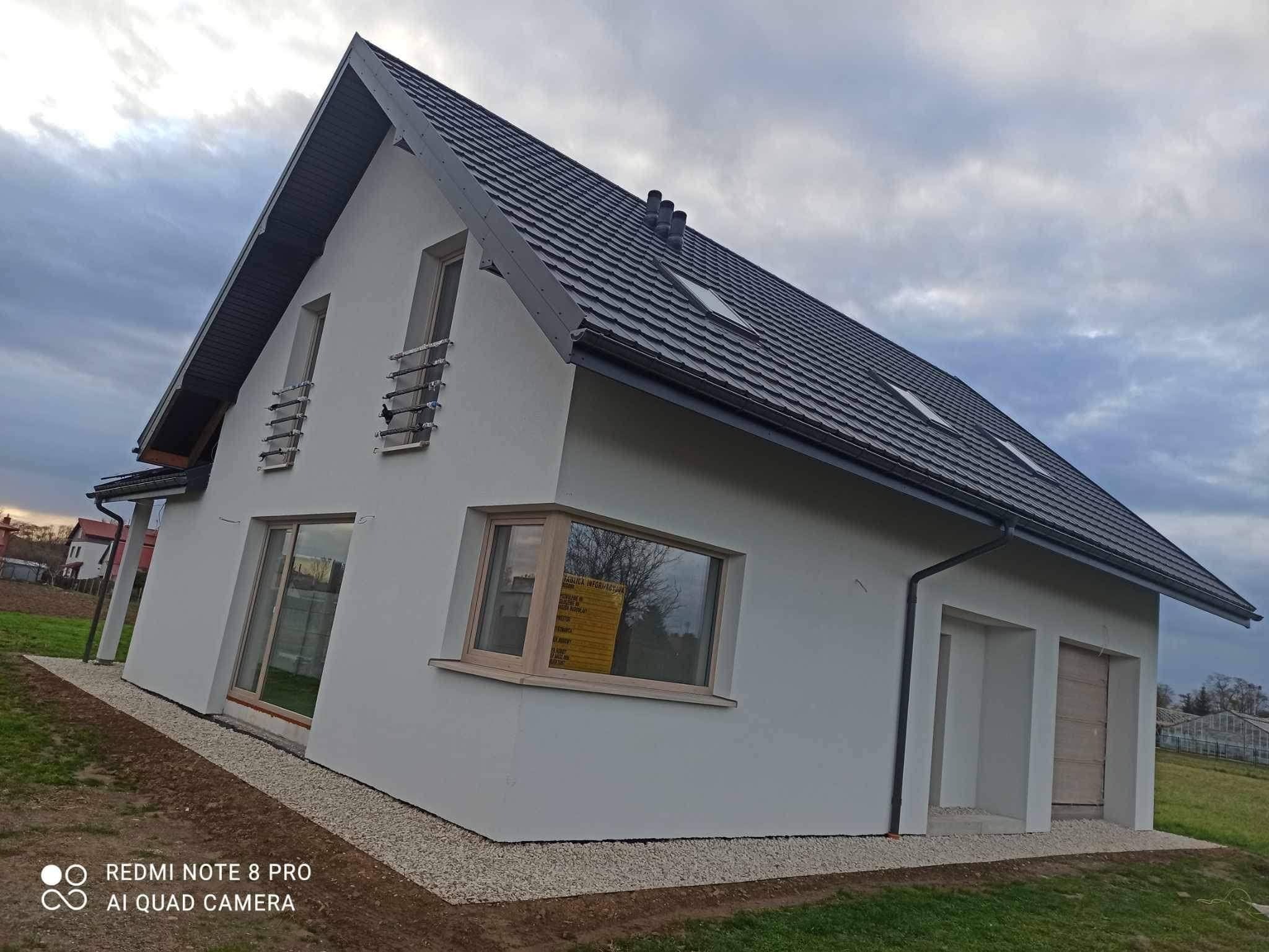 Nowoczesny dom jednorodzinny z szarą dachówką i białymi ścianami, widok z zewnątrz na tle pochmurnego nieba, z widocznymi oknami i elementami wykończenia.