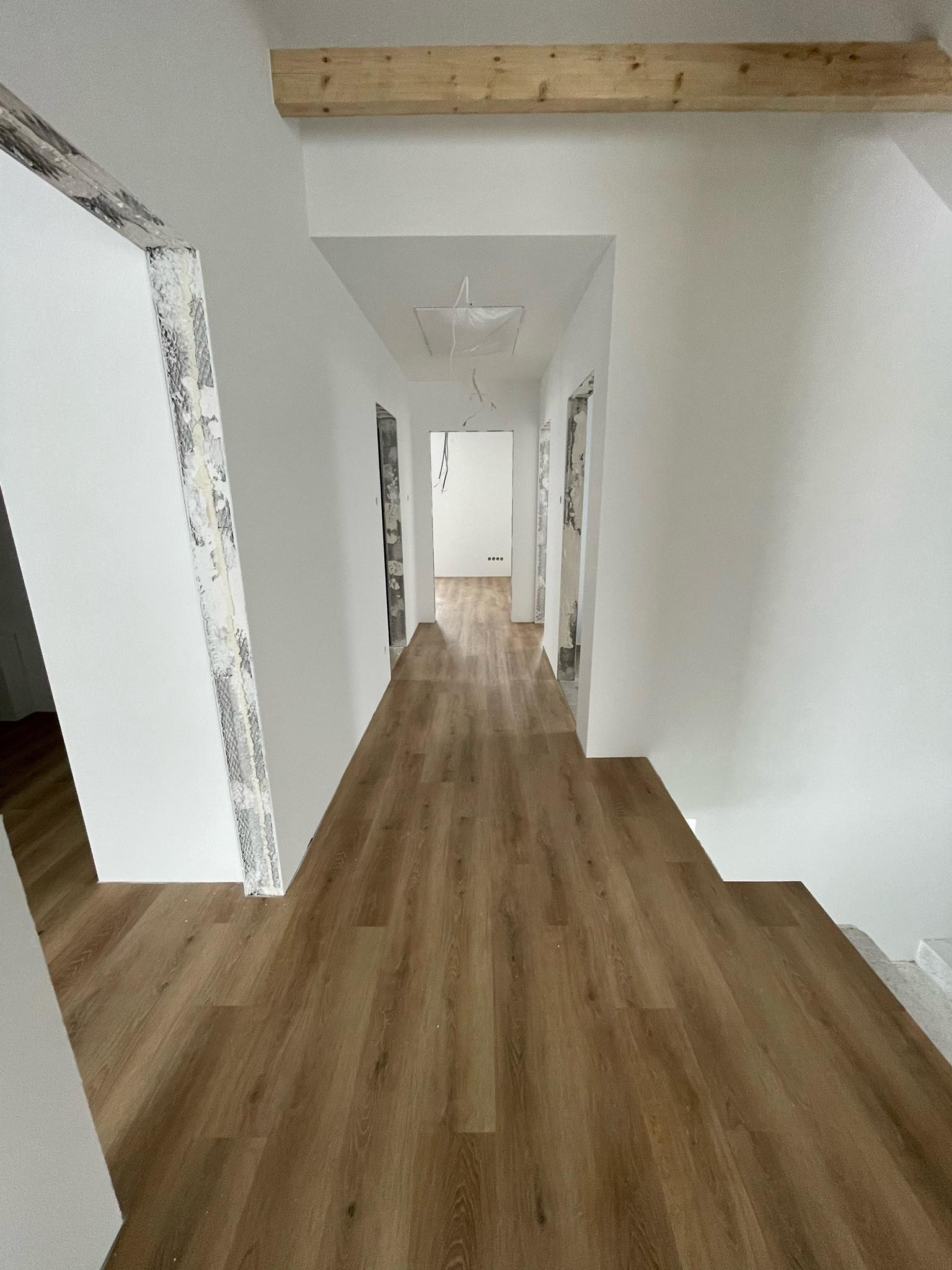 Korytarz w trakcie prac wykończeniowych: świeżo położona podłoga z paneli imitujących drewno, białe ściany, surowe wykończenie ościeżnic, widoczny element drewniany pod sufitem.
