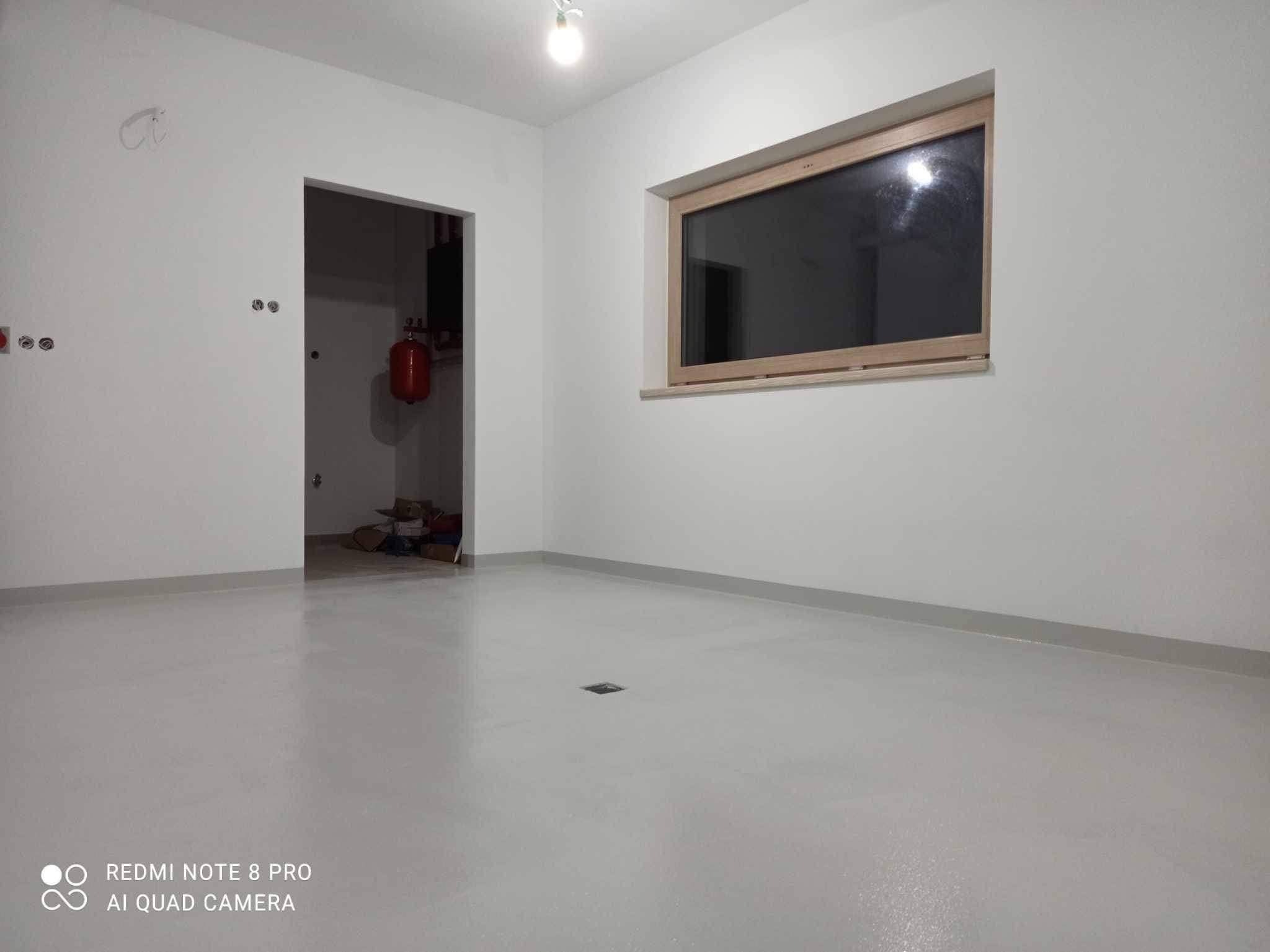 Wnętrze świeżo wykończonego pokoju z gładkimi, białymi ścianami, drewnianym oknem i szarą wylewką podłogową. Widoczny grzejnik w pomieszczeniu obok przez otwarte drzwi.