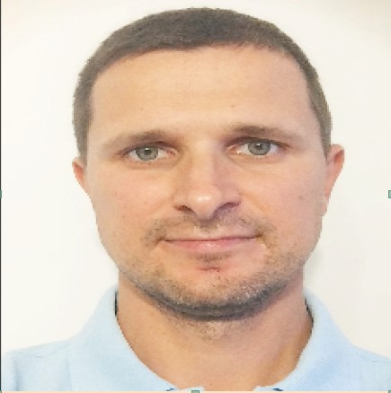 Portret mężczyzny z krótkimi włosami, niebieskimi oczami i lekkim zarostem, ubrany w jasnoniebieską koszulkę polo, na białym tle.