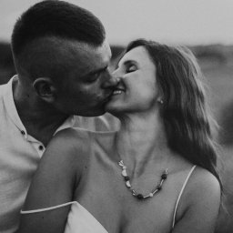 Anna Wiszowata Fotografia - Czarno-białe zbliżenie całującej się pary. Kobieta z naszyjnikiem i sukienką na ramiączkach, mężczyzna w koszuli z rozpiętym kołnierzykiem. Romantyczny moment uchwycony w plenerze.