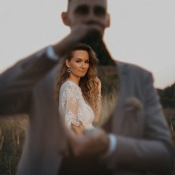 Anna Wiszowata Fotografia - Kreatywne ujęcie pary: kobieta w koronkowej sukience na pierwszym planie, mężczyzna w beżowym garniturze w tle, z efektem głębi i naturalnym oświetleniem.