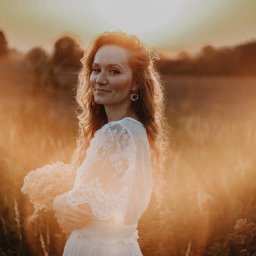 Anna Wiszowata Fotografia - Uśmiechnięta kobieta w zwiewnej sukience z koronkowymi rękawami, trzymająca bukiet na tle zachodzącego słońca, w plenerze.