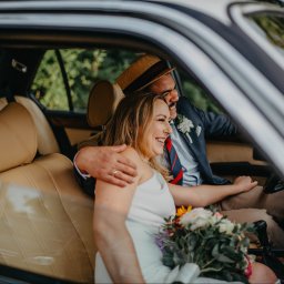 Anna Wiszowata Fotografia - Uśmiechnięta para młoda w samochodzie z bukietem. Pan młody w kapeluszu i marynarce, panna młoda w białej sukience. Jasne wnętrze auta, widok z boku przez szybę.