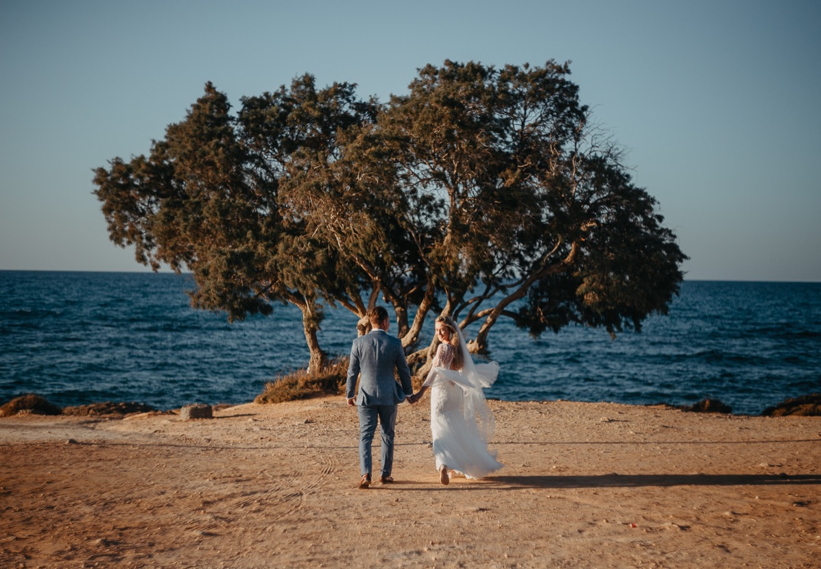 Młoda para trzymająca się za ręce, idzie w stronę morza na tle drzewa. Pan młody w niebieskim garniturze, panna młoda w białej sukni z welonem. Ujęcie z tyłu.