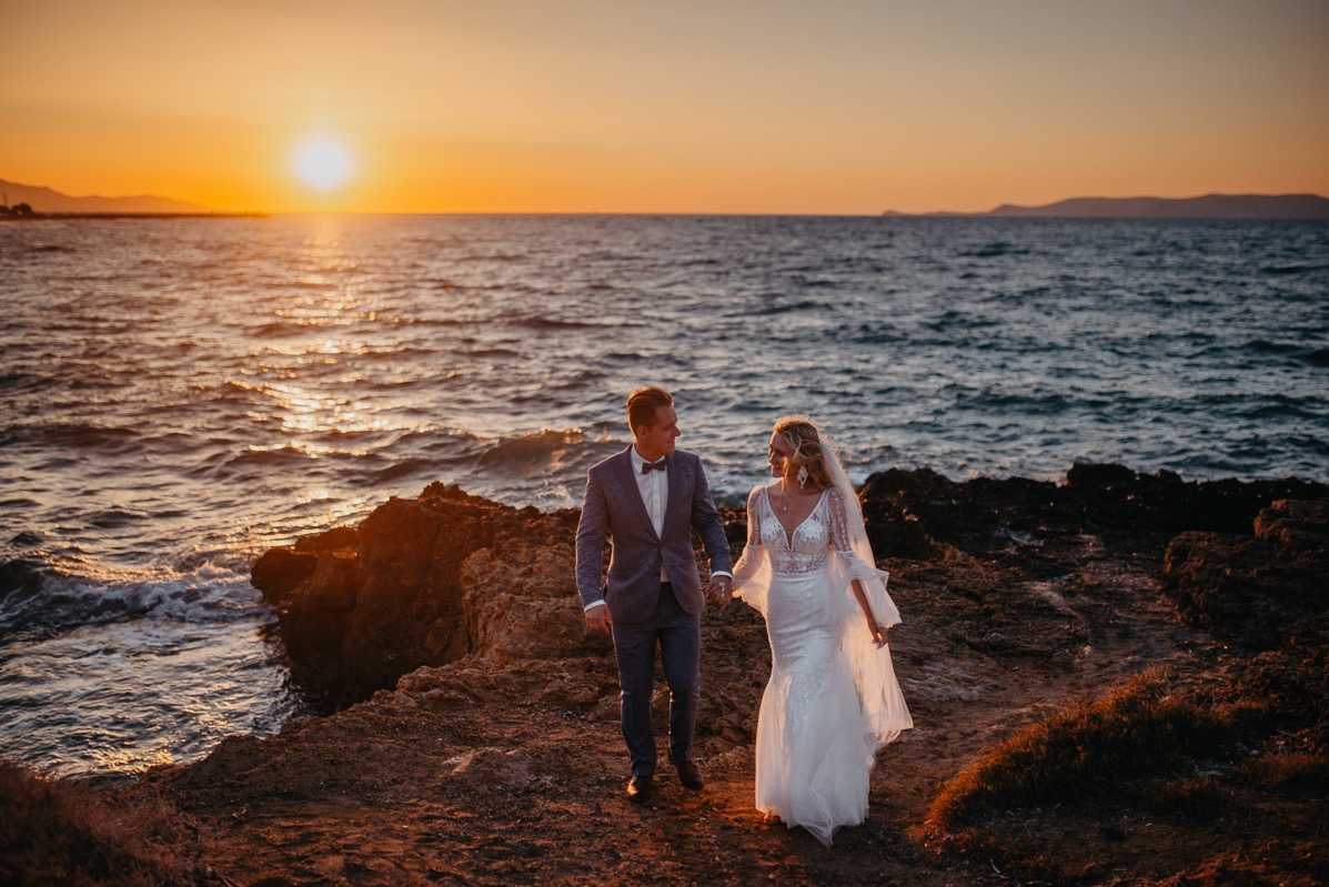 Para młoda trzymająca się za ręce na tle zachodzącego słońca nad morzem. Pan młody w granatowym garniturze, panna młoda w białej sukni z welonem.
