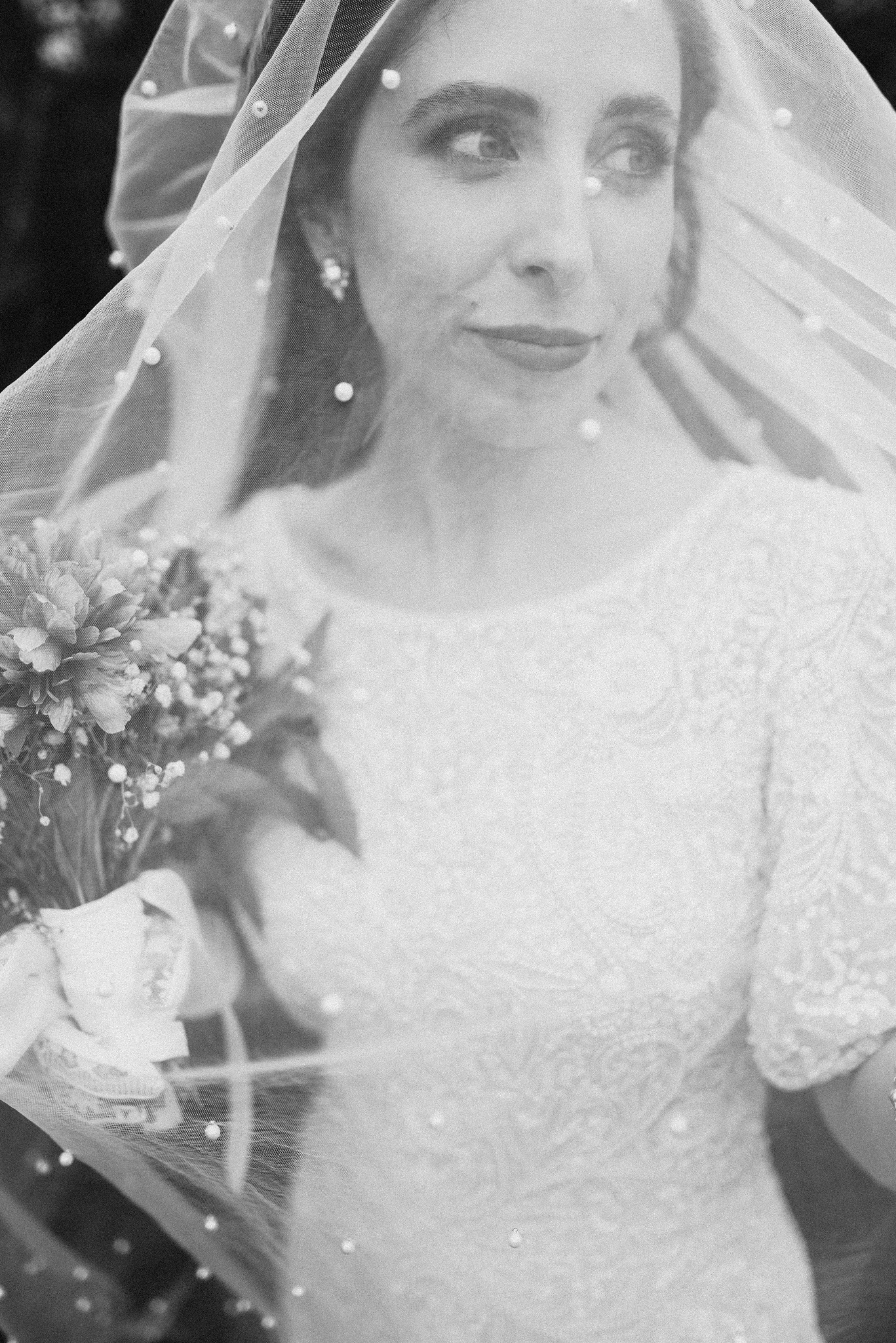 Portret panny młodej w welonie z perłami, trzymającej bukiet kwiatów, w czarno-białej tonacji, z delikatnym uśmiechem i spojrzeniem w bok, podczas sesji ślubnej.