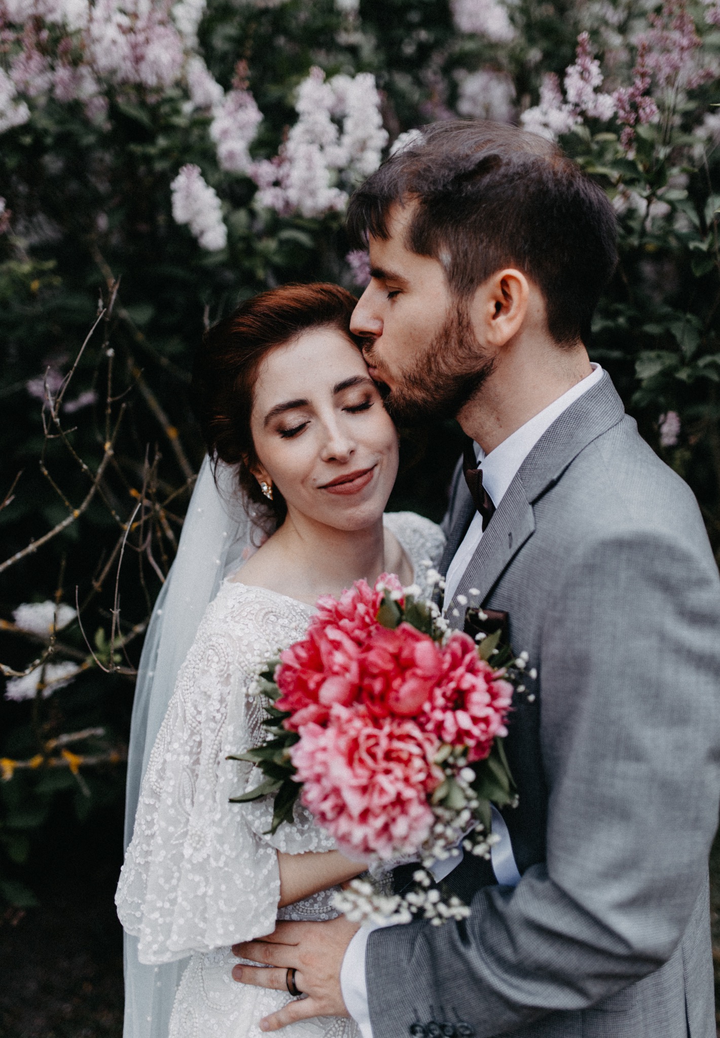 Panna młoda w koronkowej sukni ślubnej z bukietem piwonii, oblubieniec w szarym garniturze całuje ją w czoło na tle kwitnącego bzu. Romantyczna scena.