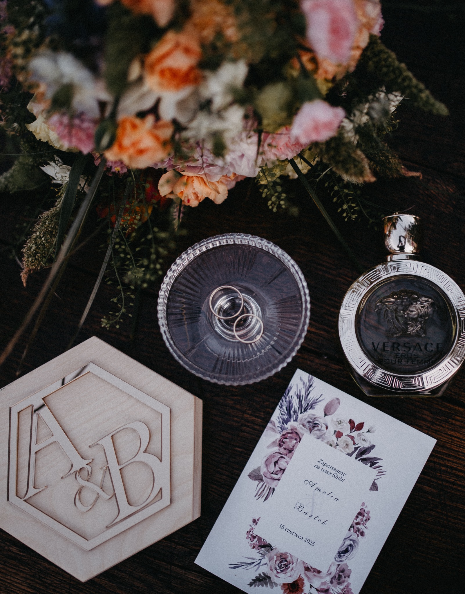 Detale ślubne: obrączki w szklanym naczyniu, zaproszenie z datą 15 czerwca 2025, drewniane pudełko z inicjałami A&B, bukiet kwiatów i flakon perfum Versace.