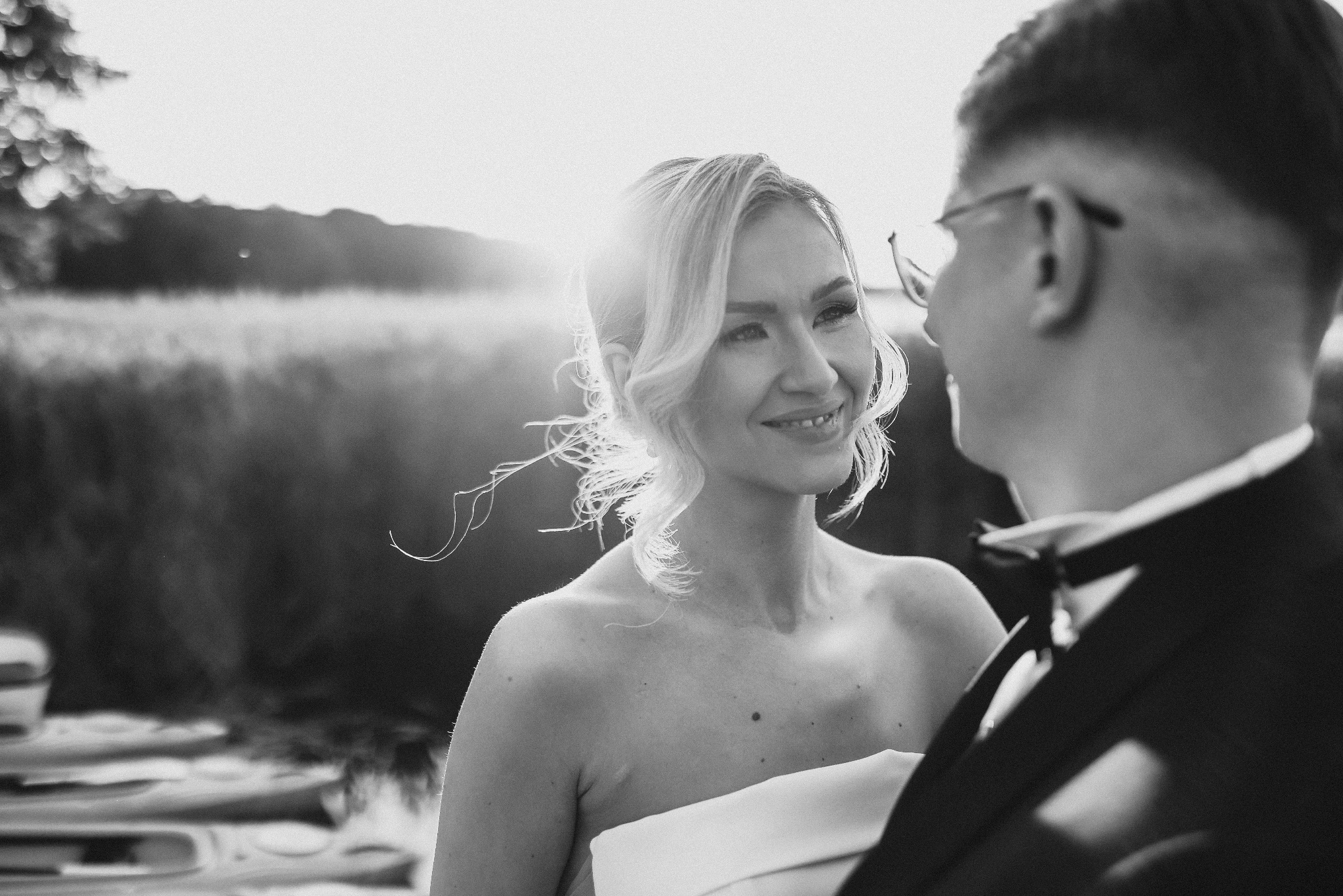 Czarno-białe zdjęcie uśmiechniętej panny młodej spoglądającej na pana młodego na tle jeziora i zachodzącego słońca.