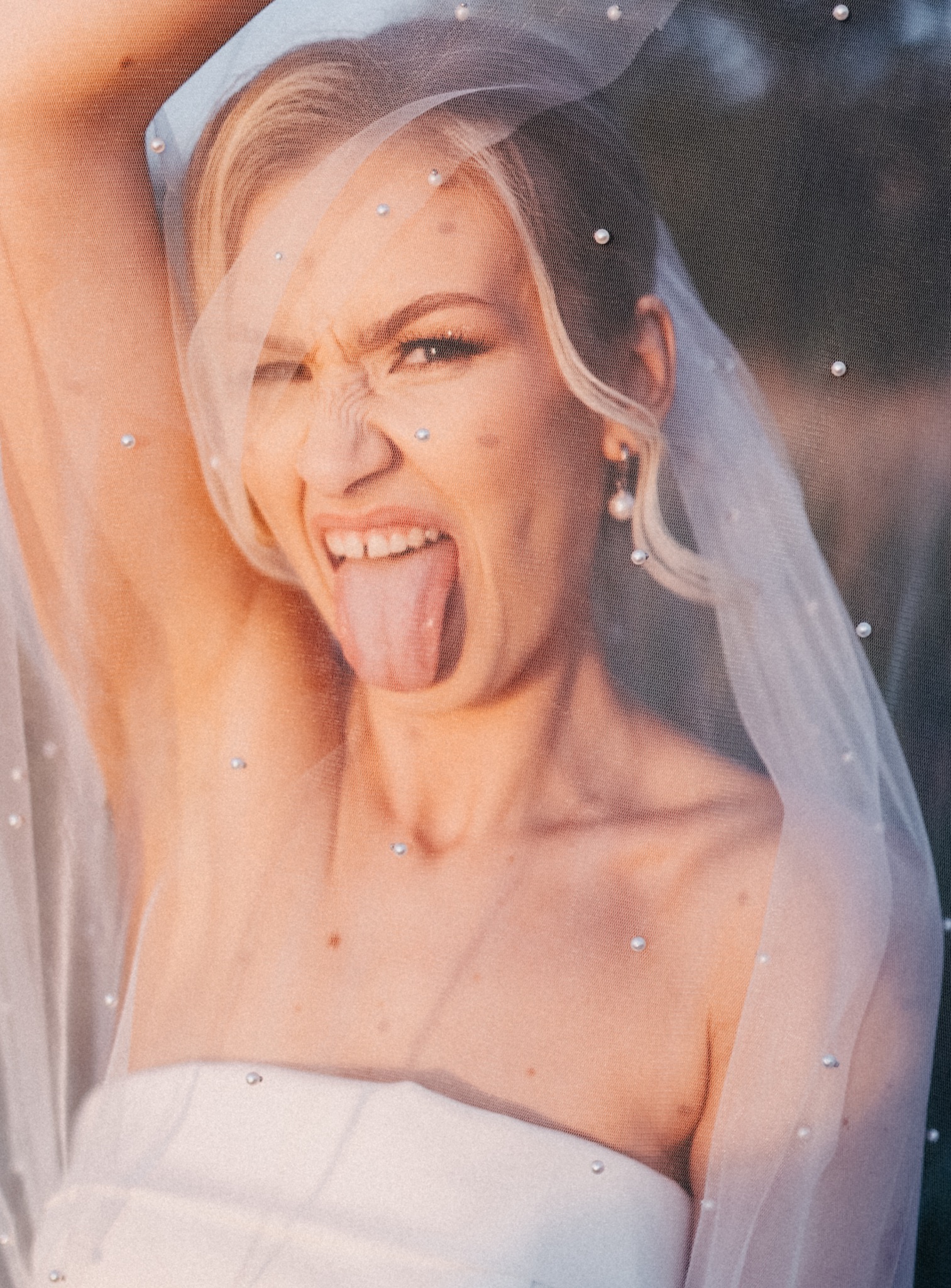 Panna młoda w białej sukni i welonie z perełkami, wystawia język i robi zabawną minę, patrząc w obiektyw. Nietypowe ujęcie w plenerze.